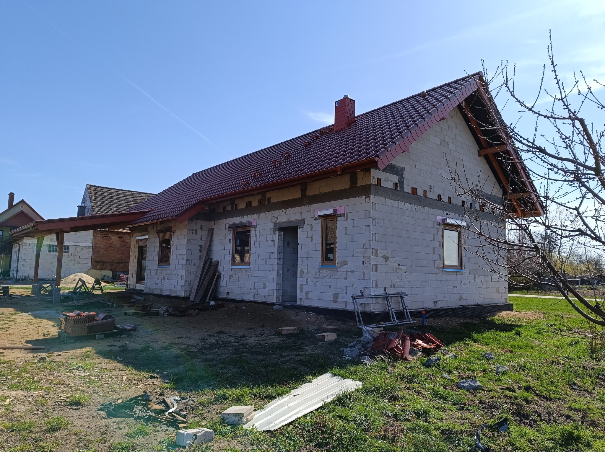 Surowy stan domu jednorodzinnego w trakcie budowy: ściany z białych bloczków, czerwona dachówka, widoczne okna i rusztowanie, trawa na pierwszym planie.