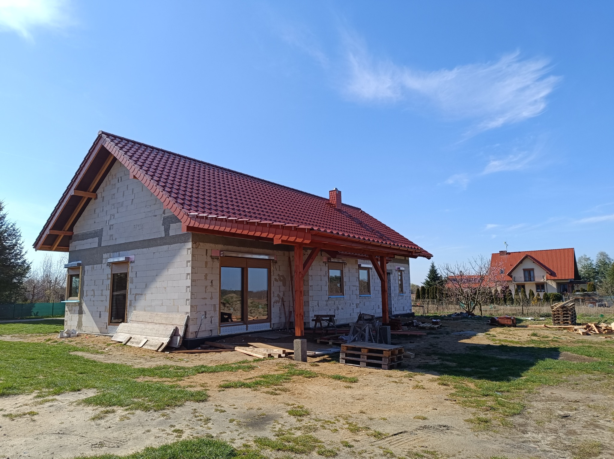 Surowy stan domu jednorodzinnego z czerwonym dachem, widoczne białe bloczki, drewniana konstrukcja zadaszenia tarasu, okna z roletami zewnętrznymi, palety i deski na trawniku, drugi dom w tle...