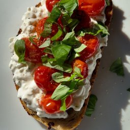 Magda Orzechowska - Kromka chleba z kremowym serem, pieczonymi pomidorkami cherry i świeżą bazylią na białym talerzu w naturalnym świetle.