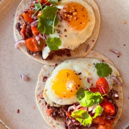 Magda Orzechowska - Dwa tacos z jajkiem sadzonym, czerwoną fasolą, pomidorami i kolendrą, posypane płatkami chili, na talerzu z ceramicznym wykończeniem.
