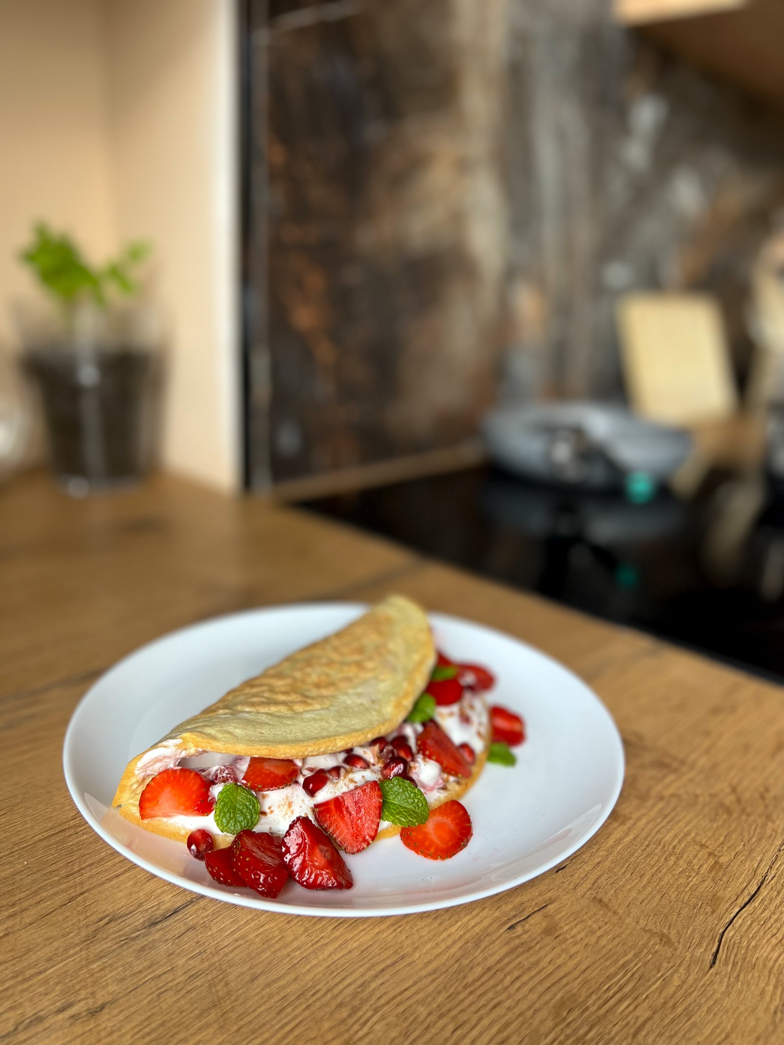 Naleśnik faszerowany bitą śmietaną, truskawkami i miętą na białym talerzu na drewnianym blacie kuchennym, w tle roślina w doniczce i płyta indukcyjna.