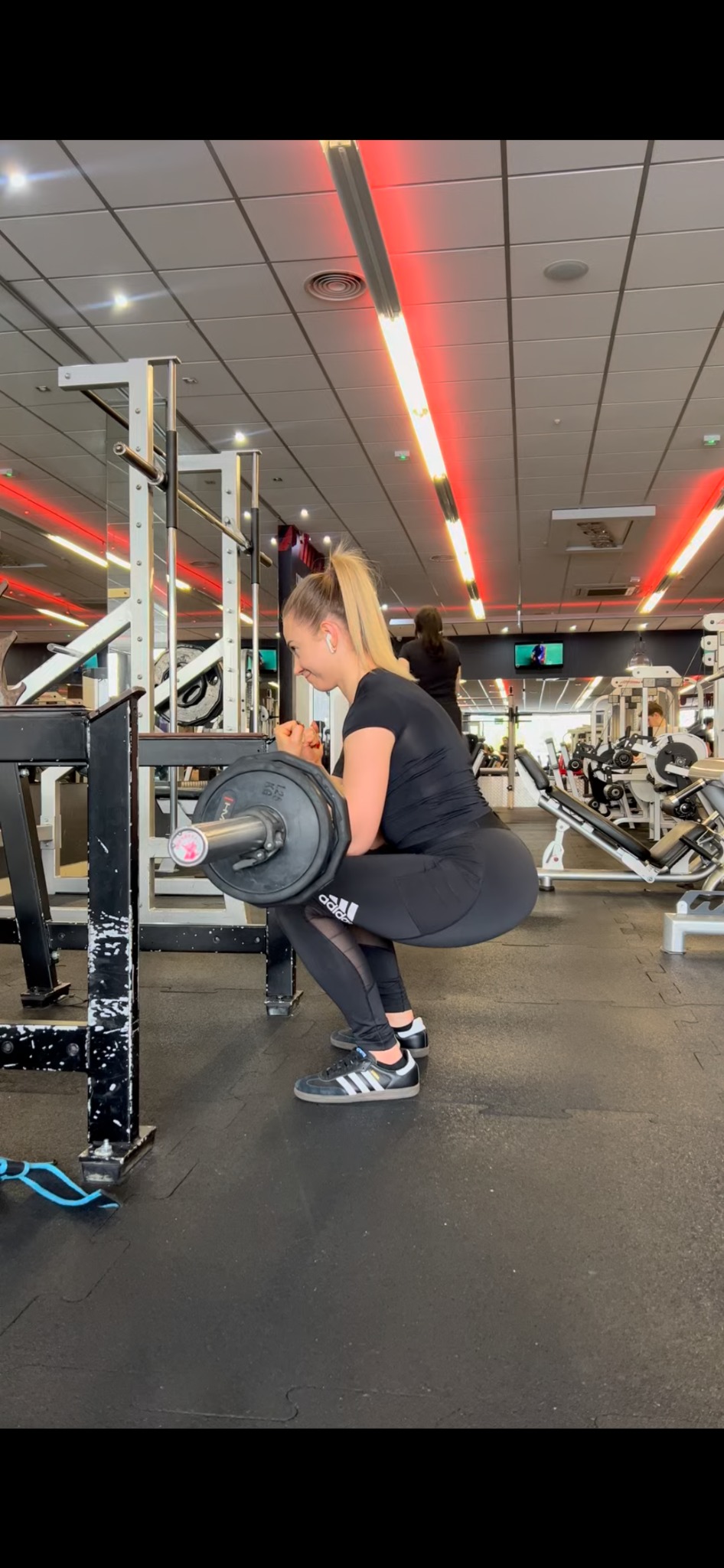 Kobieta w czarnym stroju sportowym wykonuje przysiad ze sztangą w siłowni, widoczne Adidas Samba na nogach.