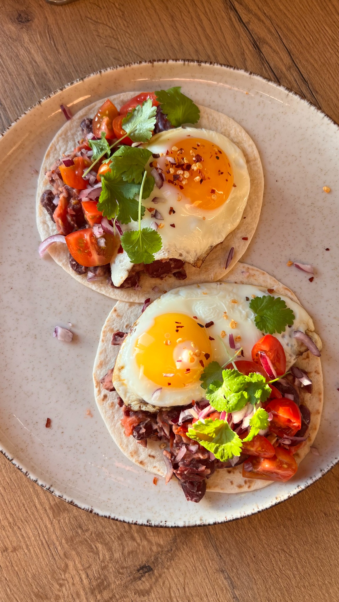 Dwa tacos z jajkiem sadzonym, czerwoną fasolą, pomidorami i kolendrą, posypane płatkami chili, na talerzu z ceramicznym wykończeniem.