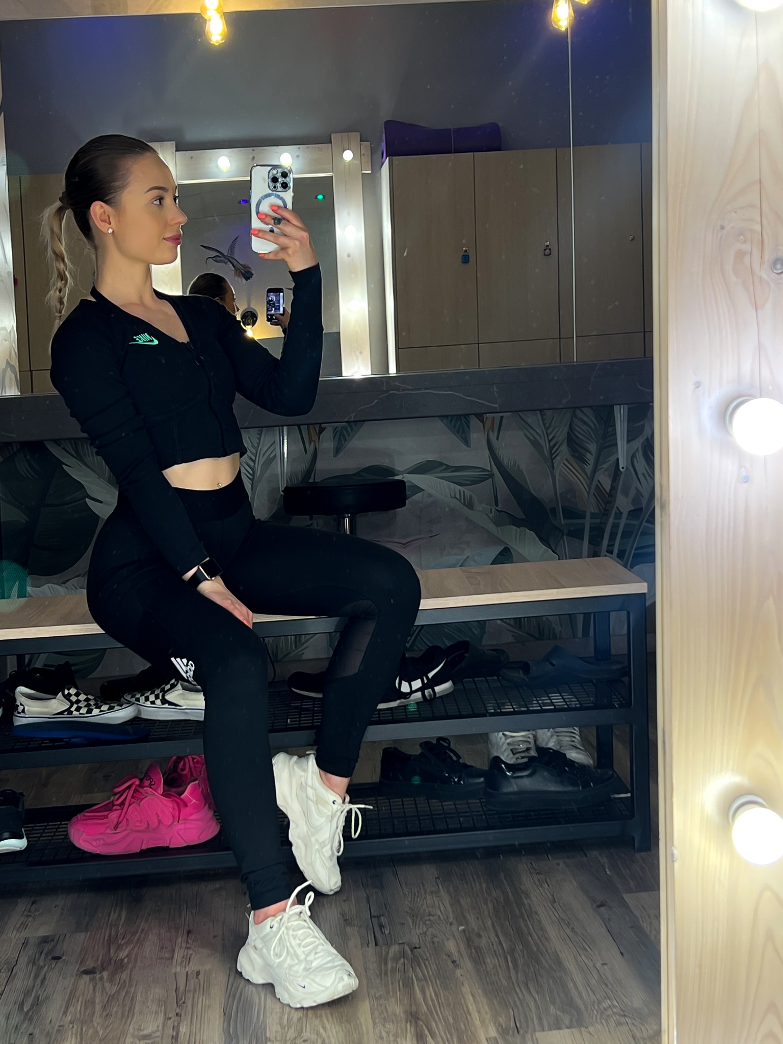 Kobieta w czarnym stroju sportowym, siedząca na ławce w szatni, robi selfie w lustrze, na półkach pod ławką widoczne różne pary butów.