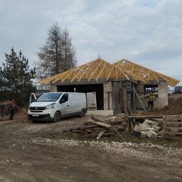 MAR-BUD - Budowa domu jednorodzinnego w trakcie wznoszenia konstrukcji dachu z drewnianych krokwi; na pierwszym planie biały samochód dostawczy firmy budowlanej, w tle drzewa i pochmurne niebo.