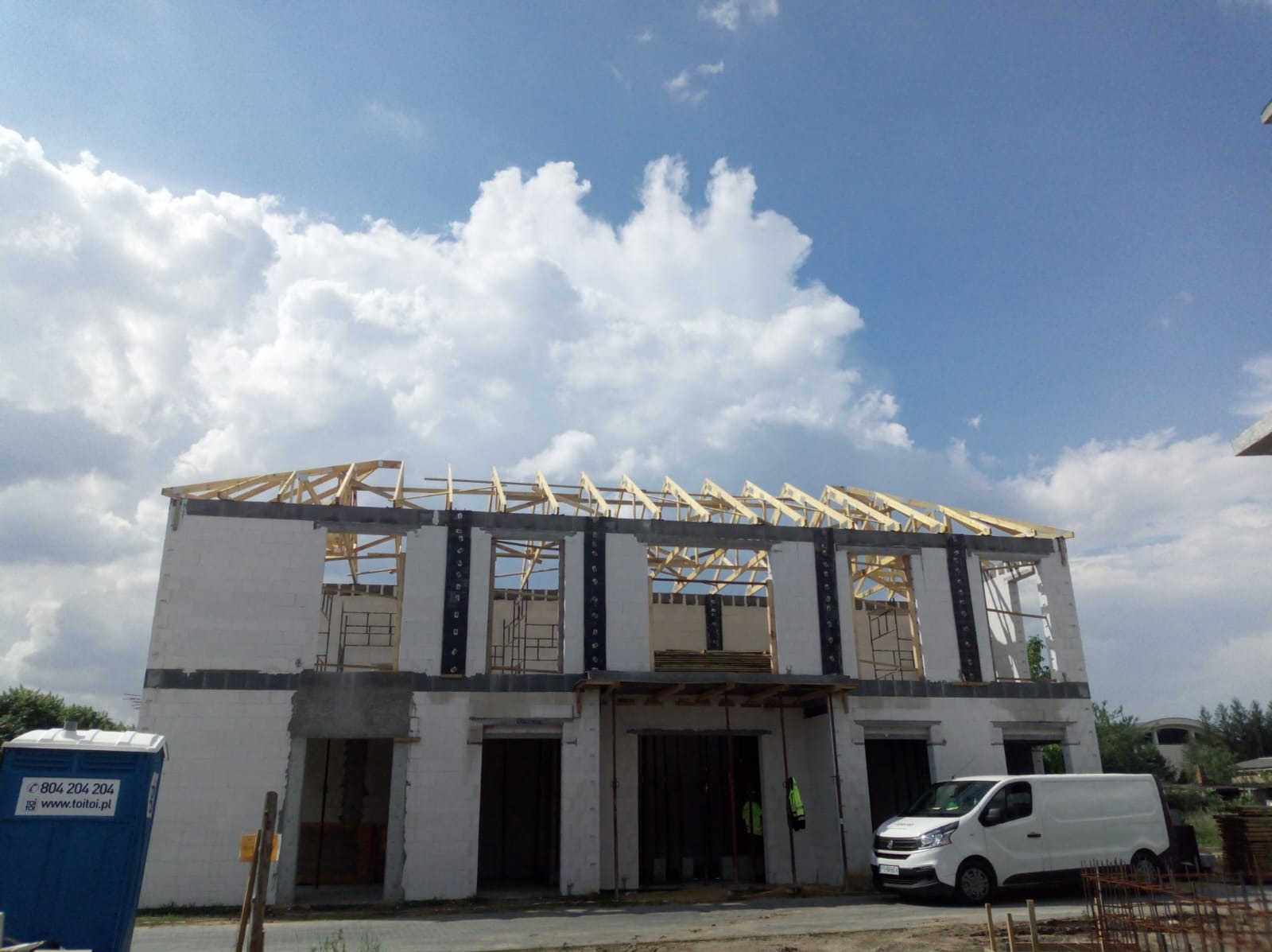 Budowa domu jednorodzinnego w stanie surowym otwartym, widoczne ściany z bloczków, drewniana konstrukcja dachu, biały samochód dostawczy i przenośna toaleta na placu budowy, pochmurne niebo w tle.