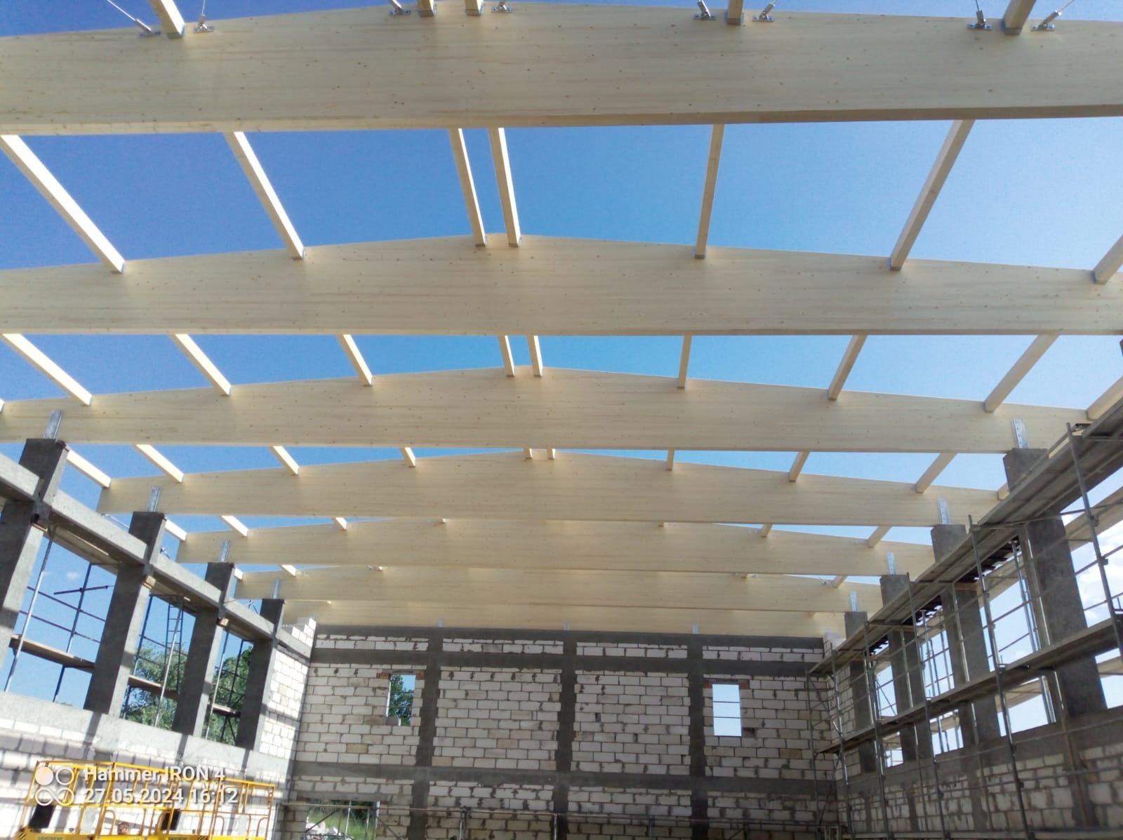 Konstrukcja dachu z jasnego drewna w surowym stanie, widok od wewnątrz budynku w trakcie budowy, błękitne niebo w tle.