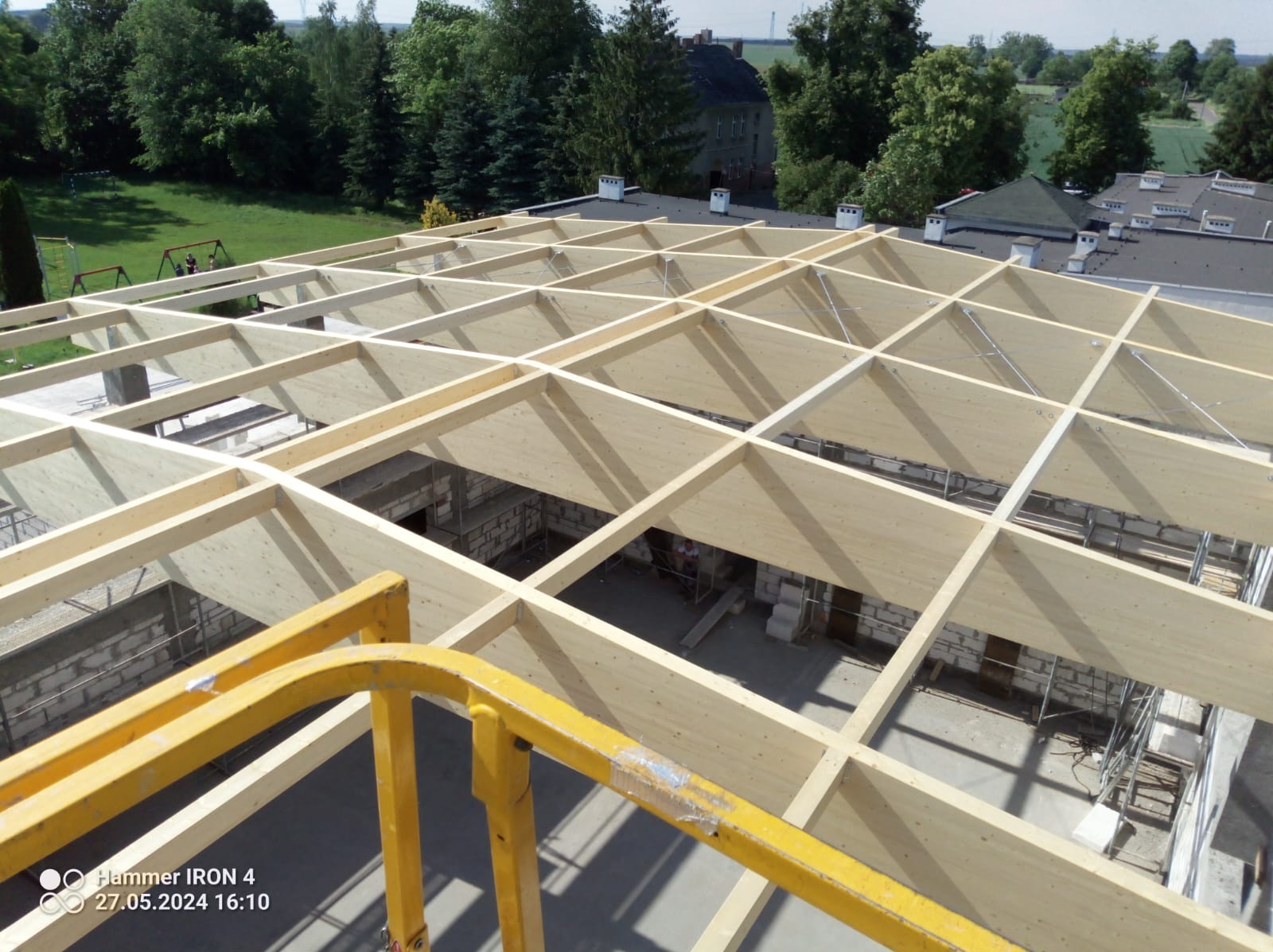 Drewniana konstrukcja dachu w trakcie budowy, widok z góry z podnośnika, jasne drewno, elementy muru z białej cegły, w tle budynki i zieleń.