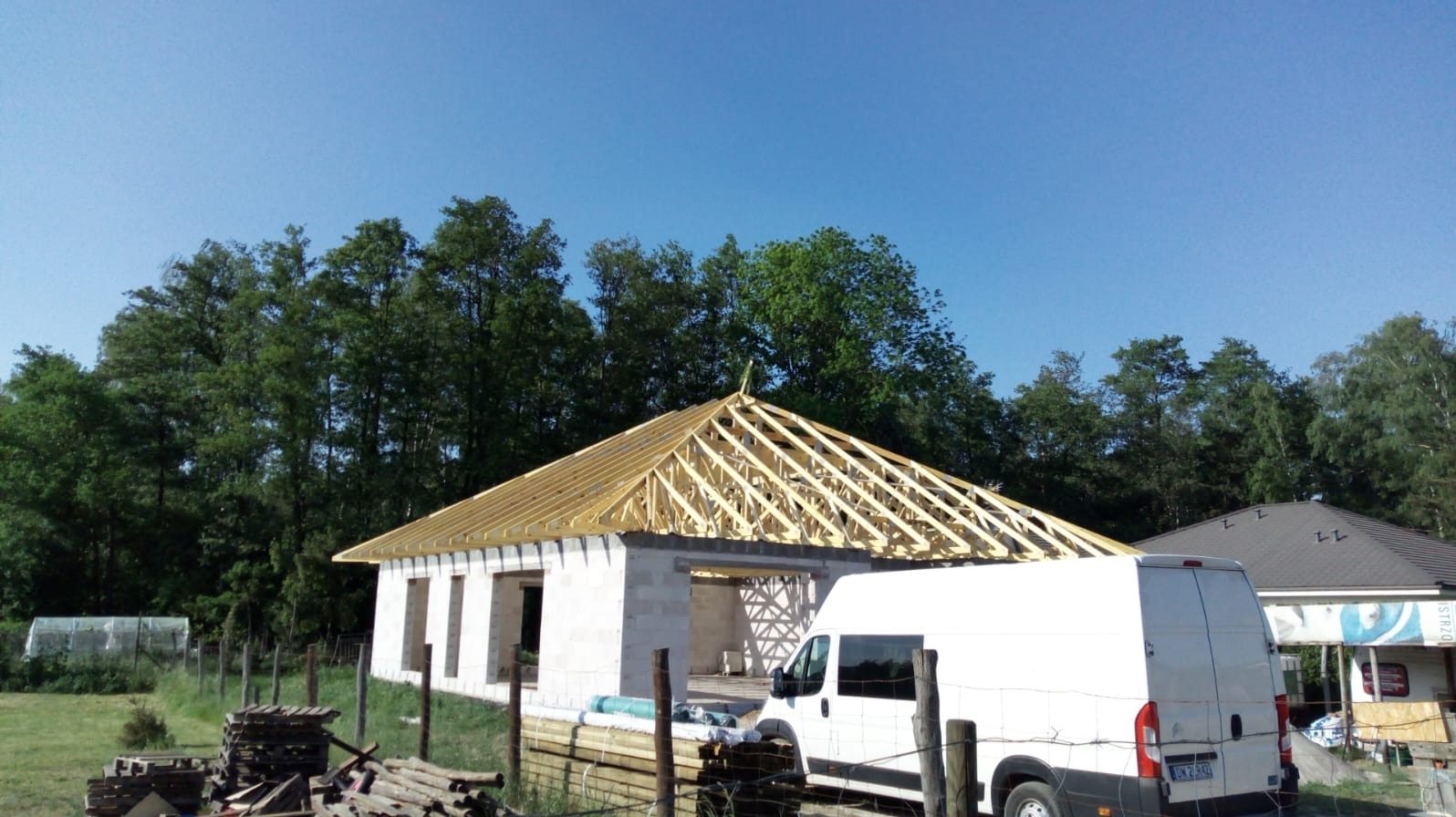 Budowa domu jednorodzinnego w trakcie: surowy budynek z bloczków, widoczna drewniana konstrukcja dachu, na pierwszym planie biały samochód dostawczy.