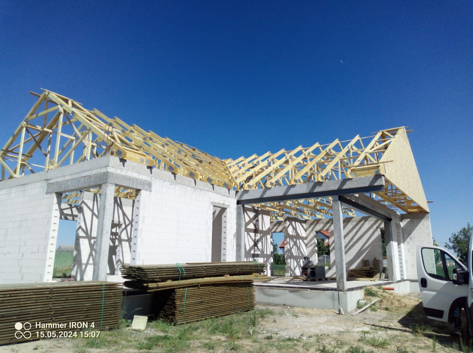 Budowa domu jednorodzinnego w stanie surowym otwartym, widoczna konstrukcja drewniana dachu z elementami deskowania, ściany z białego bloczka, ułożone drewno budowlane, błękitne niebo.