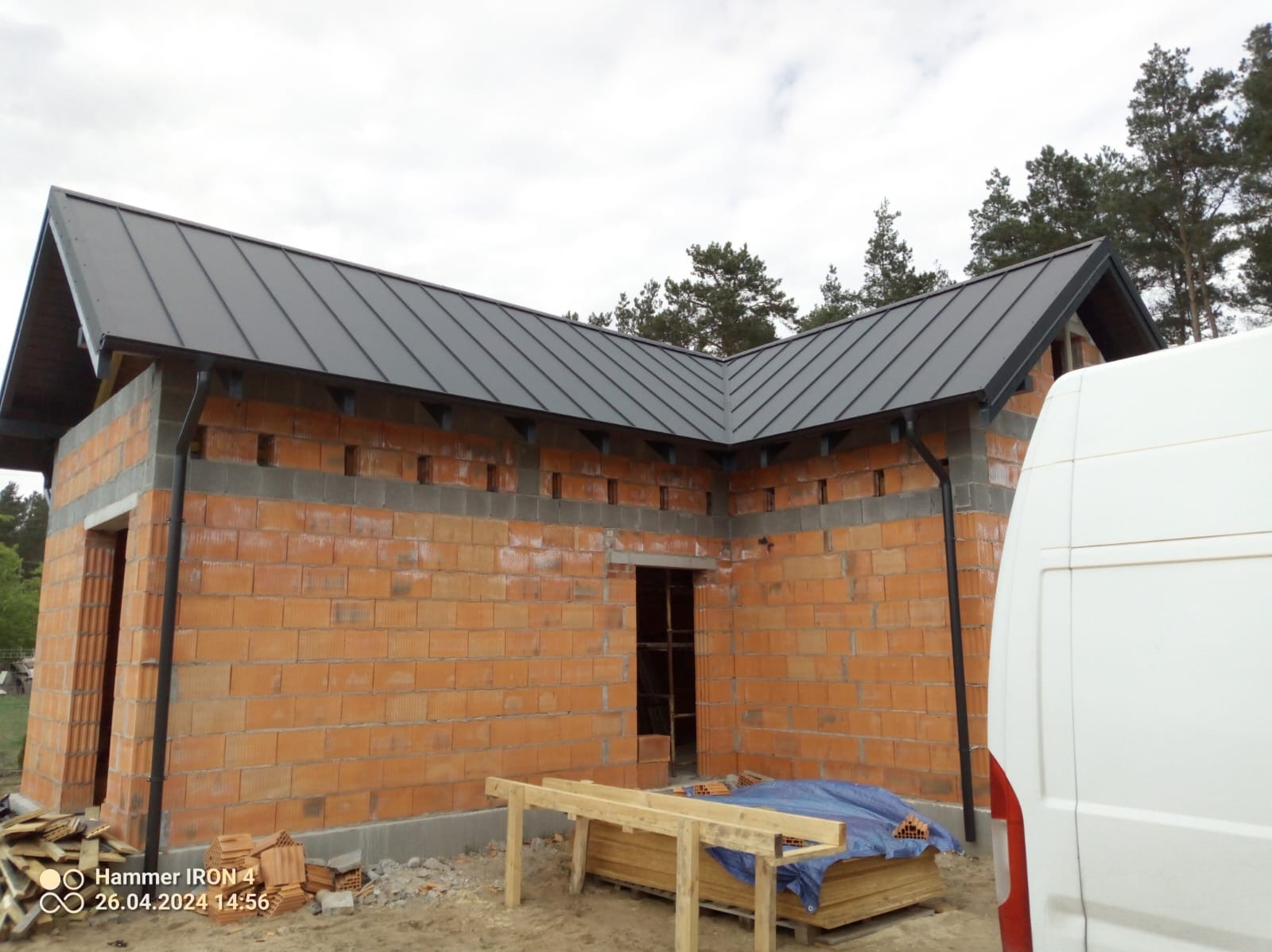 Surowy budynek w trakcie budowy z zamontowanym grafitowym dachem z blachy na rąbek stojący i orynnowaniem, widoczny fragment białego samochodu dostawczego, materiały budowlane na placu budowy.