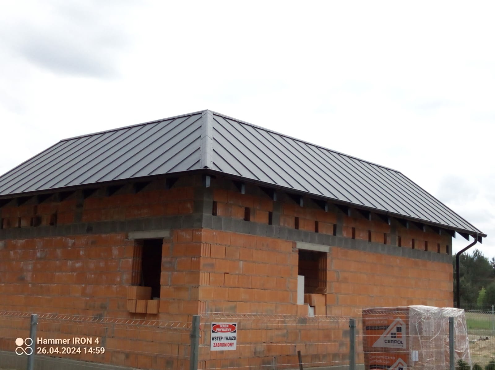 Nowoczesny, grafitowy dach z blachy na surowej bryle domu z czerwonej cegły, widoczne rynny, teren budowy zabezpieczony siatką i znakami ostrzegawczymi.
