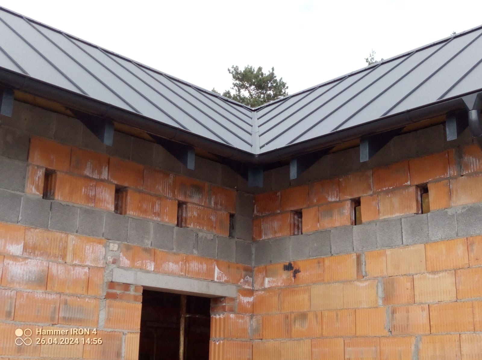 Szary dach z blachy na surowej konstrukcji budynku z cegły i betonu, widoczne wsporniki rynny i fragment rynny, w tle wierzchołek drzewa.