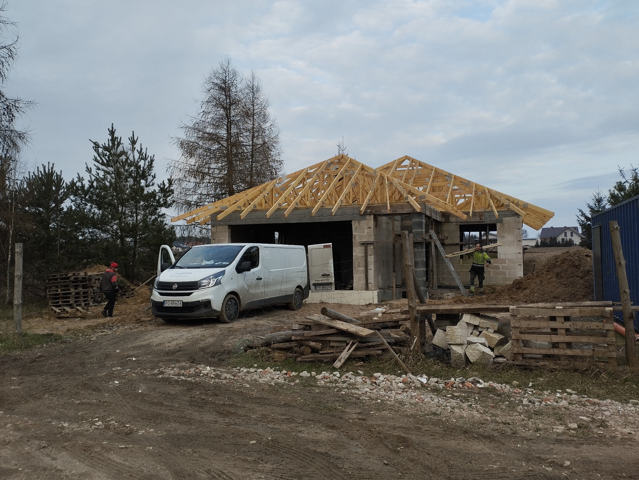 Budowa domu jednorodzinnego w trakcie wznoszenia konstrukcji dachu z drewnianych krokwi; na pierwszym planie biały samochód dostawczy firmy budowlanej, w tle drzewa i pochmurne niebo.