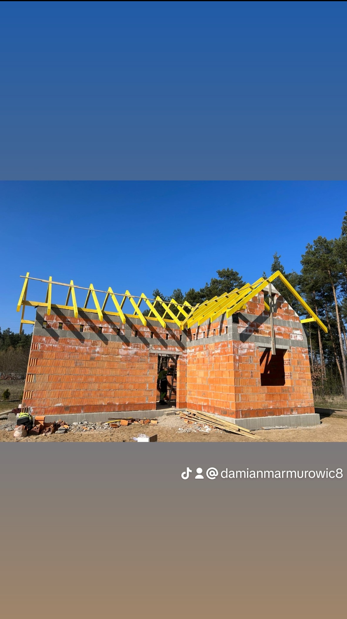 Wznoszenie murów z czerwonej cegły i montaż żółtej konstrukcji więźby dachowej na budowie domu jednorodzinnego w słoneczny dzień.