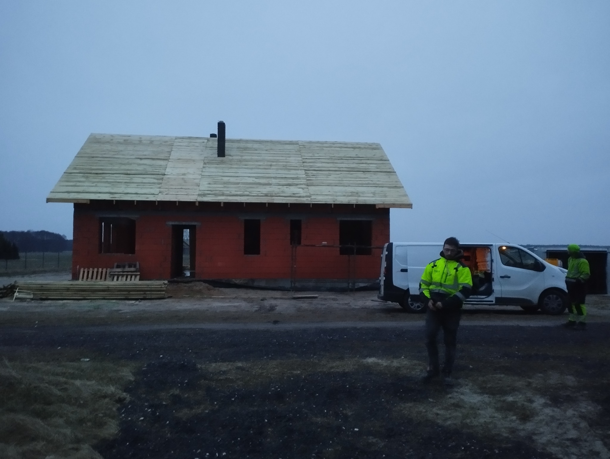 Budowa domu jednorodzinnego w Witkowie w trakcie prac dekarskich, widoczny częściowo pokryty dach drewniany i pracownicy w odblaskowych ubraniach przy samochodzie dostawczym.