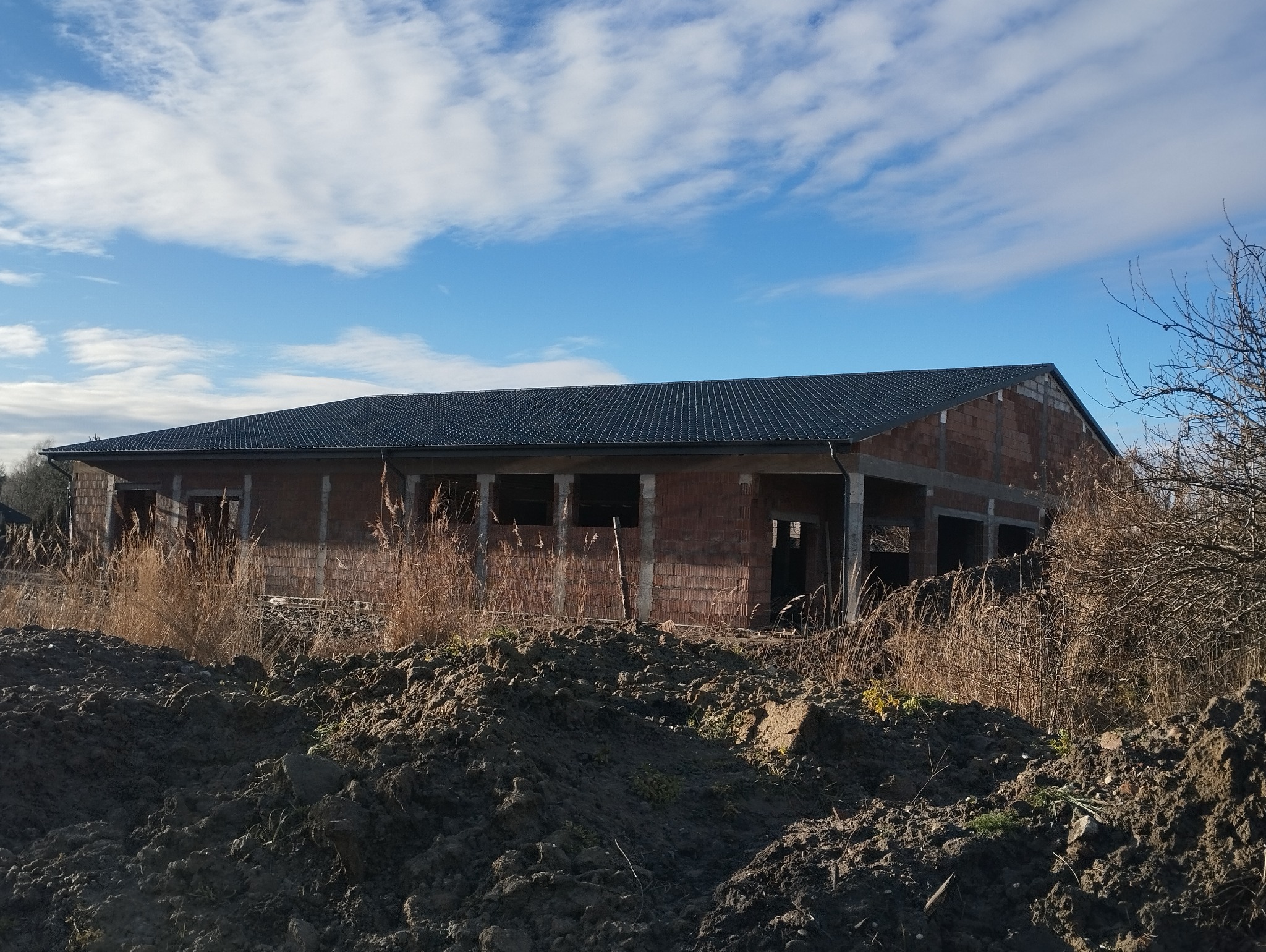 Nowy budynek w trakcie budowy z ciemnym dachem z dachówki, widoczne mury z pustaków, ziemia i trawa na pierwszym planie, błękitne niebo z chmurami w tle.