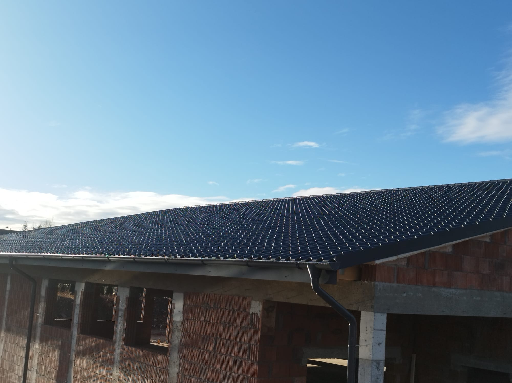 Widok z góry na nowy, czarny dach z blachy trapezowej na budynku w stanie surowym otwartym, z zamontowanymi rynnami i błękitnym niebem w tle.