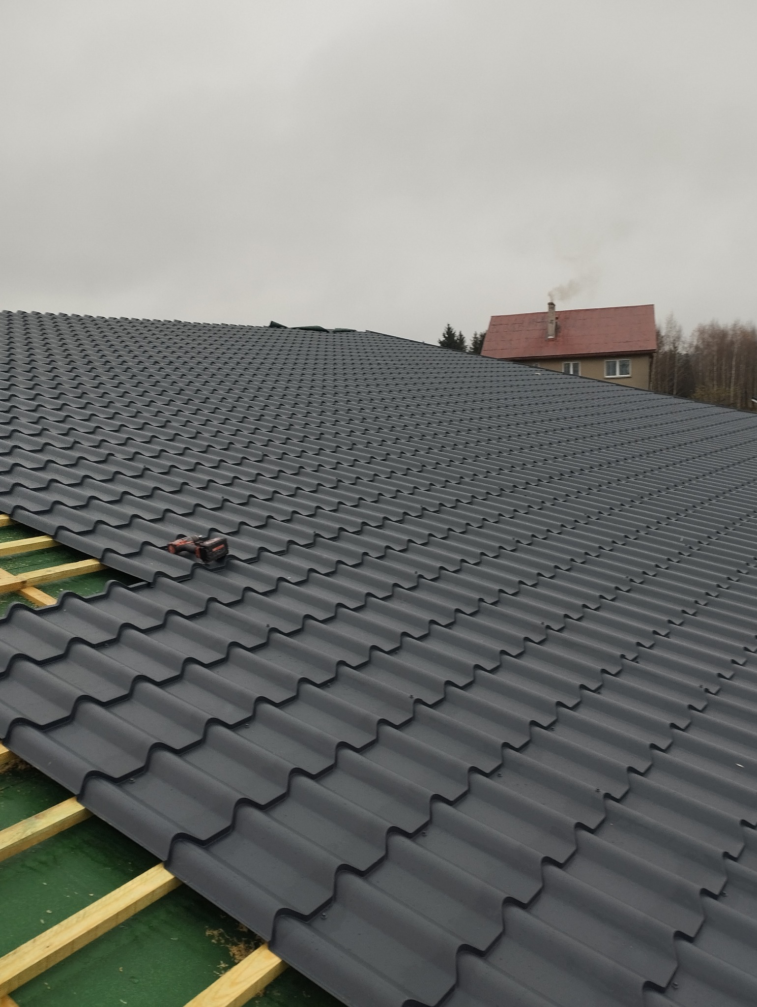 Montaż grafitowej blachodachówki na drewnianej konstrukcji dachu, w tle dom z czerwonym dachem i kominem, pochmurne niebo.