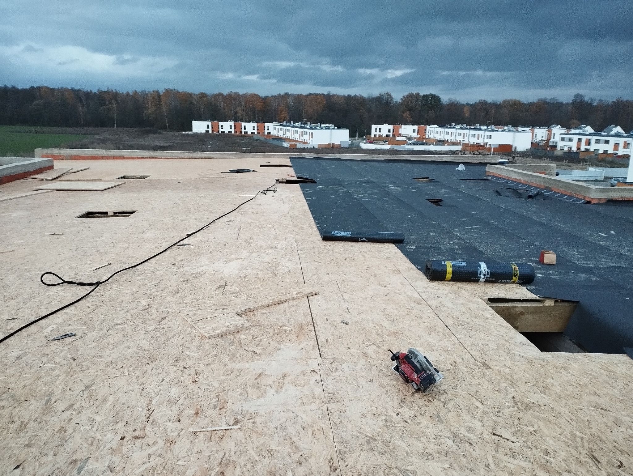 Dach w trakcie krycia: widok na poszycie z płyt OSB, częściowo pokryte czarną membraną dachową. W tle zabudowa szeregowa i pochmurne niebo.