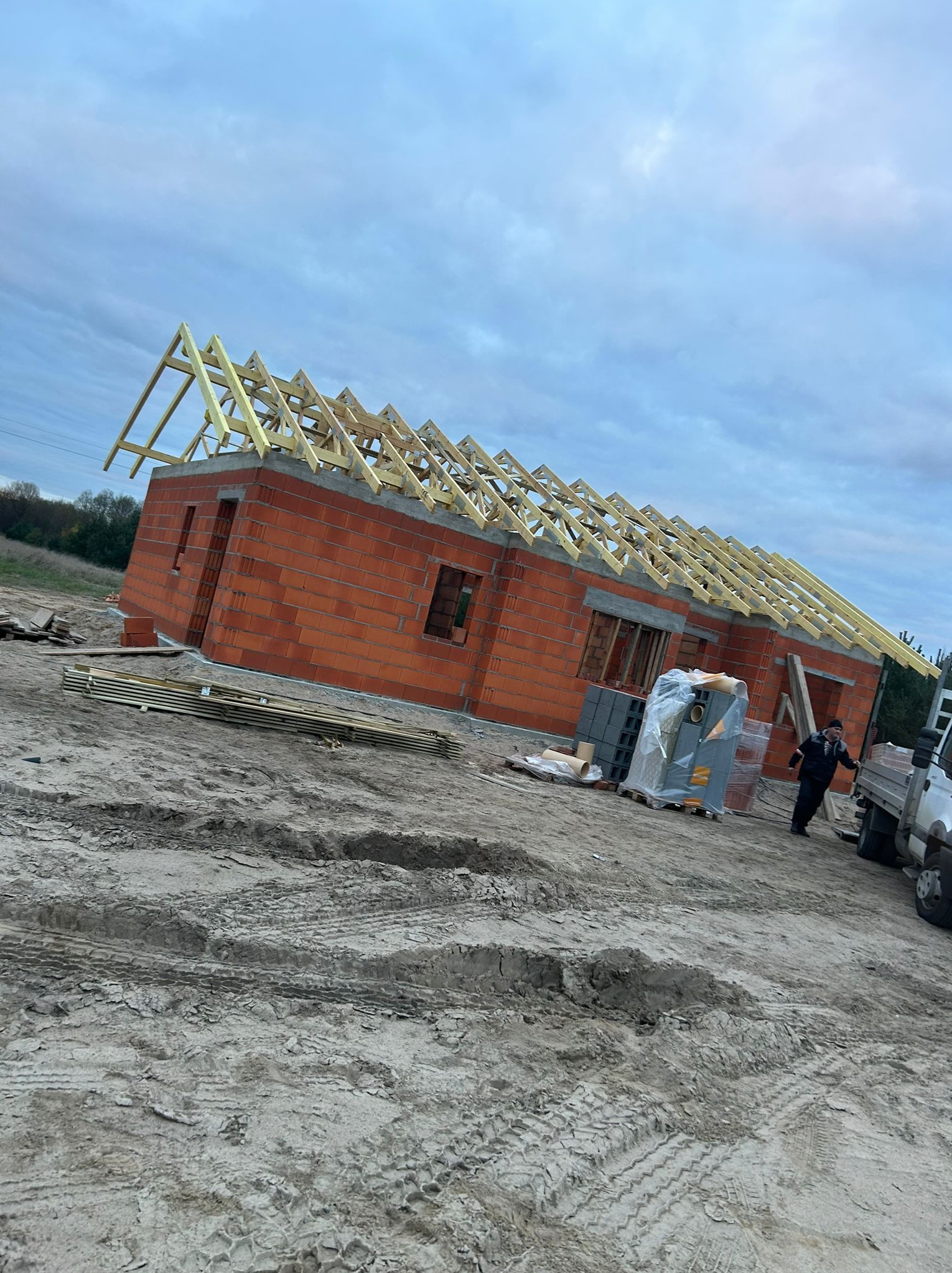 Budowa domu jednorodzinnego z czerwonej cegły: widoczne ściany, drewniana konstrukcja dachu w trakcie montażu, materiały budowlane na paletach i pracownik obok samochodu dostawczego na placu budowy.