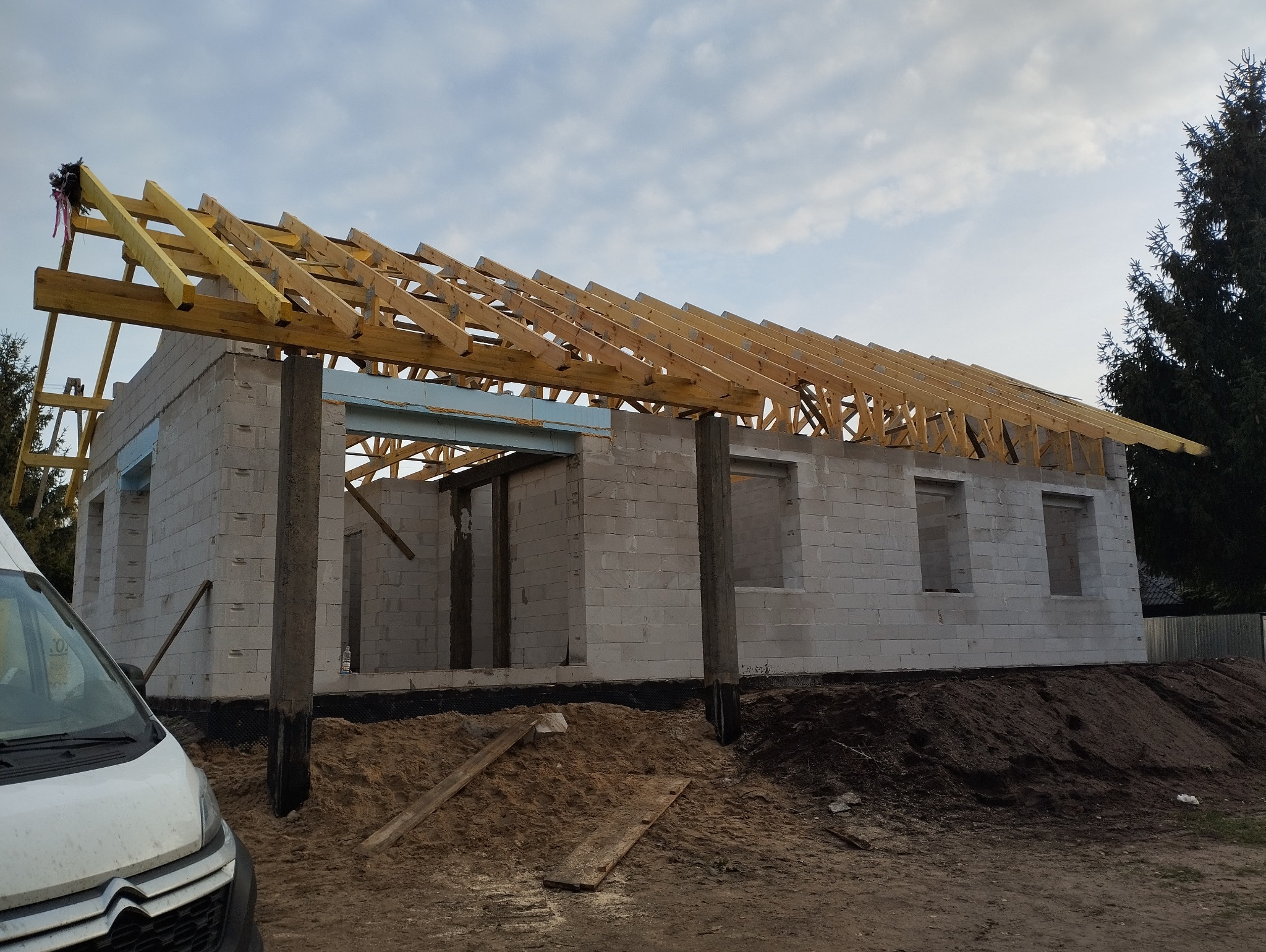 Wznoszony budynek z białych bloczków z widoczną drewnianą konstrukcją dachu, z boku zaparkowany biały samochód dostawczy.