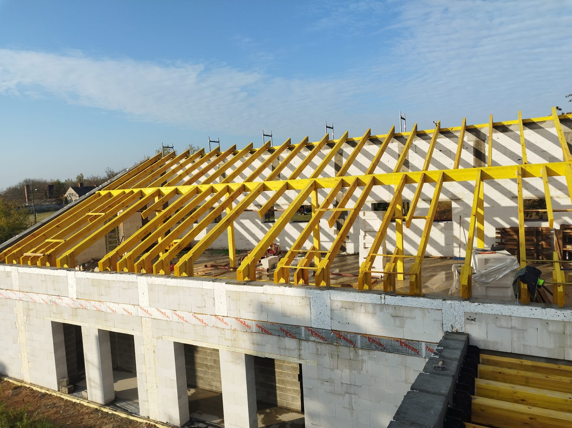 Wznoszenie konstrukcji więźby dachowej z żółtego drewna na budynku z białych bloczków, widok z góry, błękitne niebo w tle.