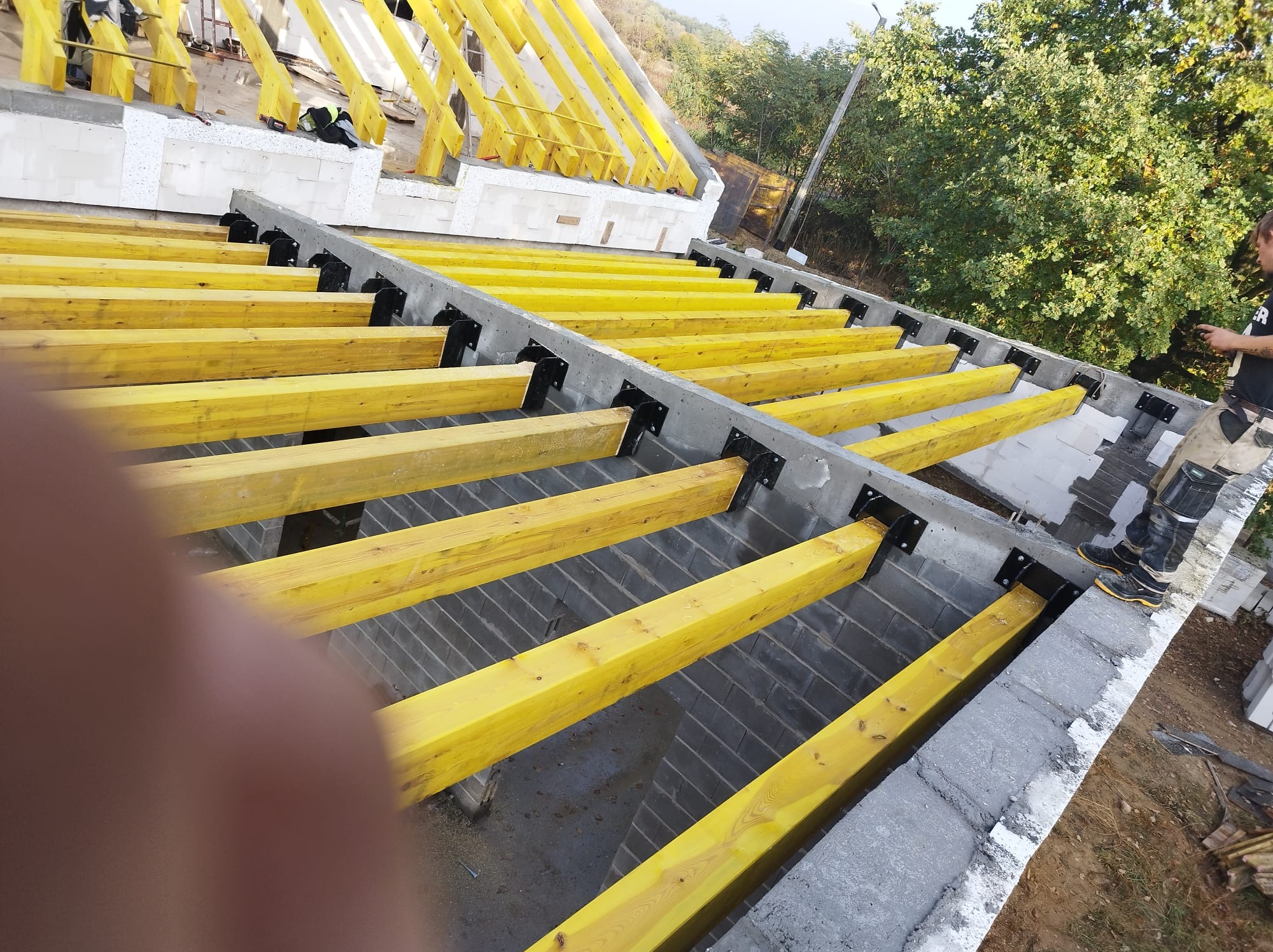 Konstrukcja dachu w trakcie budowy: żółte belki drewniane mocowane do betonowych ścian z użyciem czarnych łączników, widoczny fragment sylwetki dekarza i otoczenie budynku.