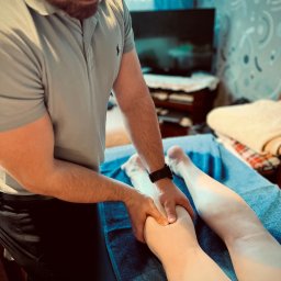 Krzysztof Dowolski Fizjoterapia - Mężczyzna w okularach, w Bytomiu, wykonuje masaż nóg pacjentki leżącej na niebieskim ręczniku na stole do masażu. W tle widoczny telewizor i wzorzysta tapeta.