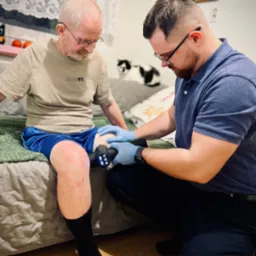 Mężczyzna w niebieskich rękawiczkach używa pistoletu do masażu na kolanie starszego mężczyzny siedzącego na łóżku w pokoju, w tle widoczny kot i dekoracje.