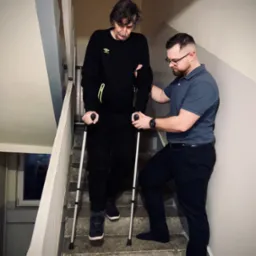 Mężczyzna z laskami schodzi po schodach, wspierany przez drugiego mężczyznę w okularach i granatowej koszulce polo.