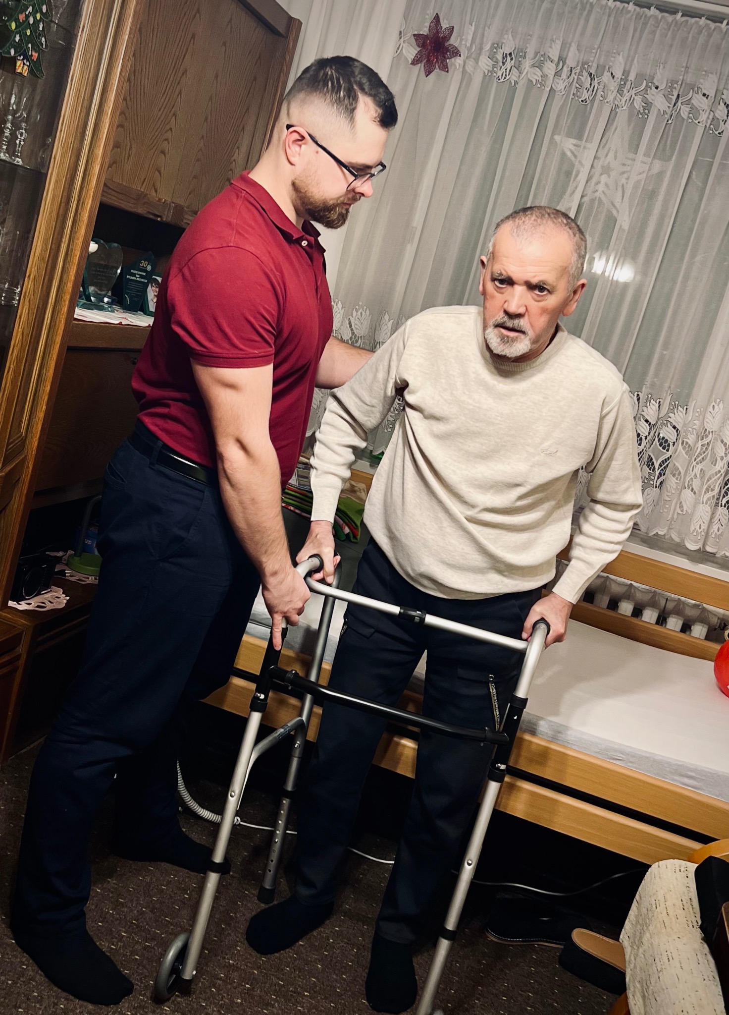 Młody mężczyzna w okularach pomaga starszemu mężczyźnie używać chodzika w pomieszczeniu z drewnianymi meblami i firankami.