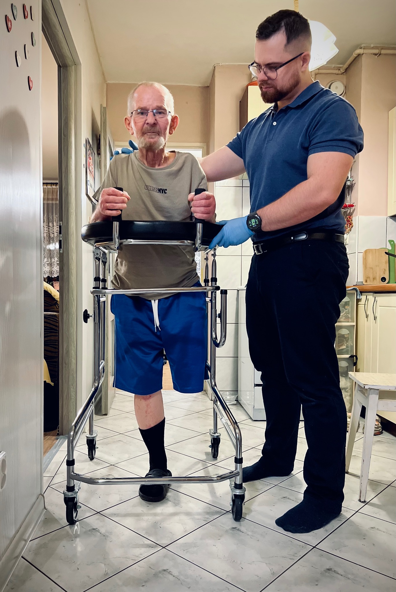 Starszy mężczyzna z chodzikiem rehabilitacyjnym, wspierany przez mężczyznę w niebieskiej koszulce polo i rękawiczkach, w domowym wnętrzu.