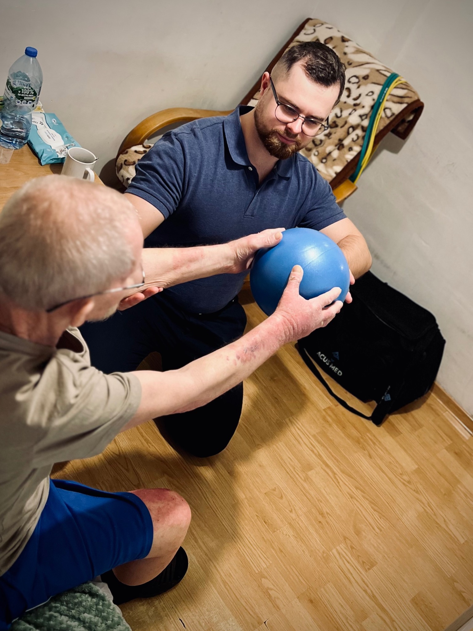 Fizjoterapeuta pomaga starszemu mężczyźnie w ćwiczeniach z niebieską piłką rehabilitacyjną w pomieszczeniu z drewnianą podłogą, widoczny worek medyczny Acus Med.