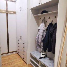 LauMaMebel Volha Lauraniuk - Biała szafa wnękowa z siedziskiem i wieszakami, uzupełniona o kurtki i czapkę, z widoczną podłogą imitującą drewno.