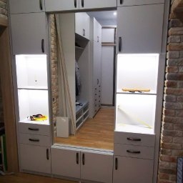 LauMaMebel Volha Lauraniuk - Szara zabudowa meblowa z lustrzanymi drzwiami przesuwnymi, wnęki z oświetleniem LED, widoczny fragment ściany z cegły i podłoga imitująca drewno. W odbiciu lustrzanym widoczna dalsza część garderoby.