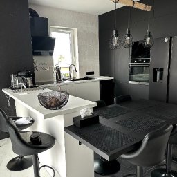 LauMaMebel Volha Lauraniuk - Nowoczesna kuchnia w czerni i bieli z wyspą i stołem barowym, oświetlona wiszącymi lampami z drewnianym elementem, z widocznym sprzętem AGD i dekoracyjnymi detalami.