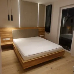 Drewniane łóżko z tapicerowanym zagłówkiem, oświetleniem LED i szafkami nocnymi wbudowanymi w ramę. Widoczny grzejnik i okno z roletą. Całość w nowoczesnym stylu.