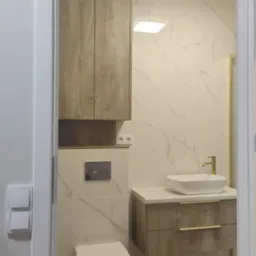 Nowoczesna łazienka z drewnianymi szafkami i umywalką nablatową. Białe marmurowe płytki, złota armatura i oświetlenie LED tworzą elegancki wystrój.