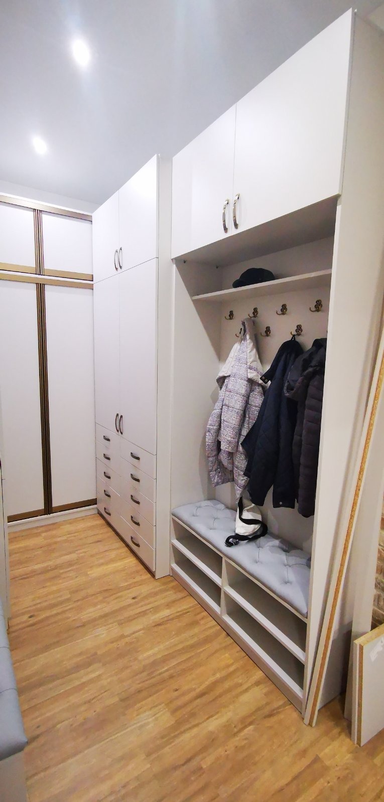 Biała szafa wnękowa z siedziskiem i wieszakami, uzupełniona o kurtki i czapkę, z widoczną podłogą imitującą drewno.