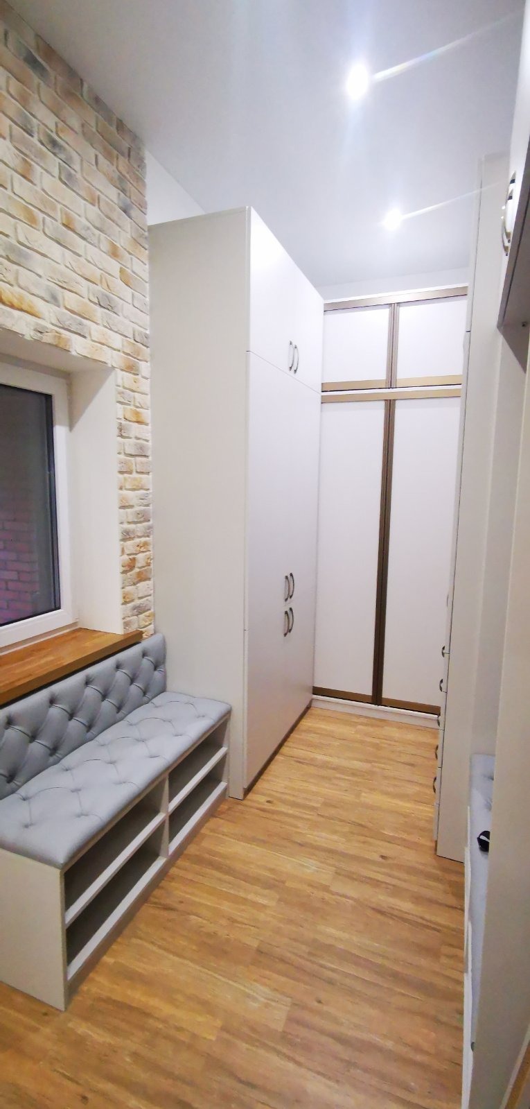 Wnętrze z zabudową: białe szafy sięgające sufitu, siedzisko z szarej tapicerki z guzikami pod oknem z ceglaną ścianą, drewniana podłoga.