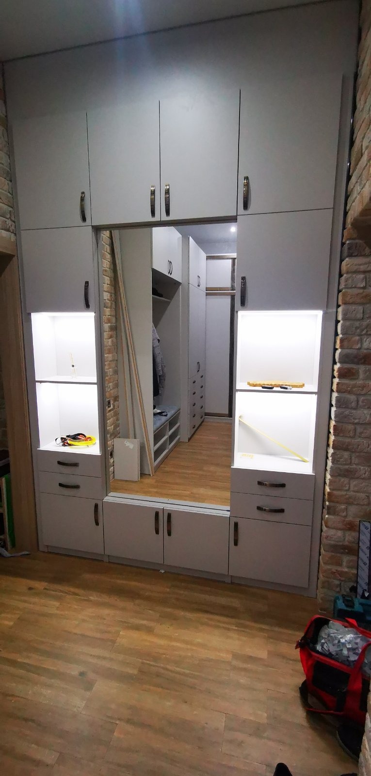 Szara zabudowa meblowa z lustrzanymi drzwiami przesuwnymi, wnęki z oświetleniem LED, widoczny fragment ściany z cegły i podłoga imitująca drewno. W odbiciu lustrzanym widoczna dalsza część garderoby.