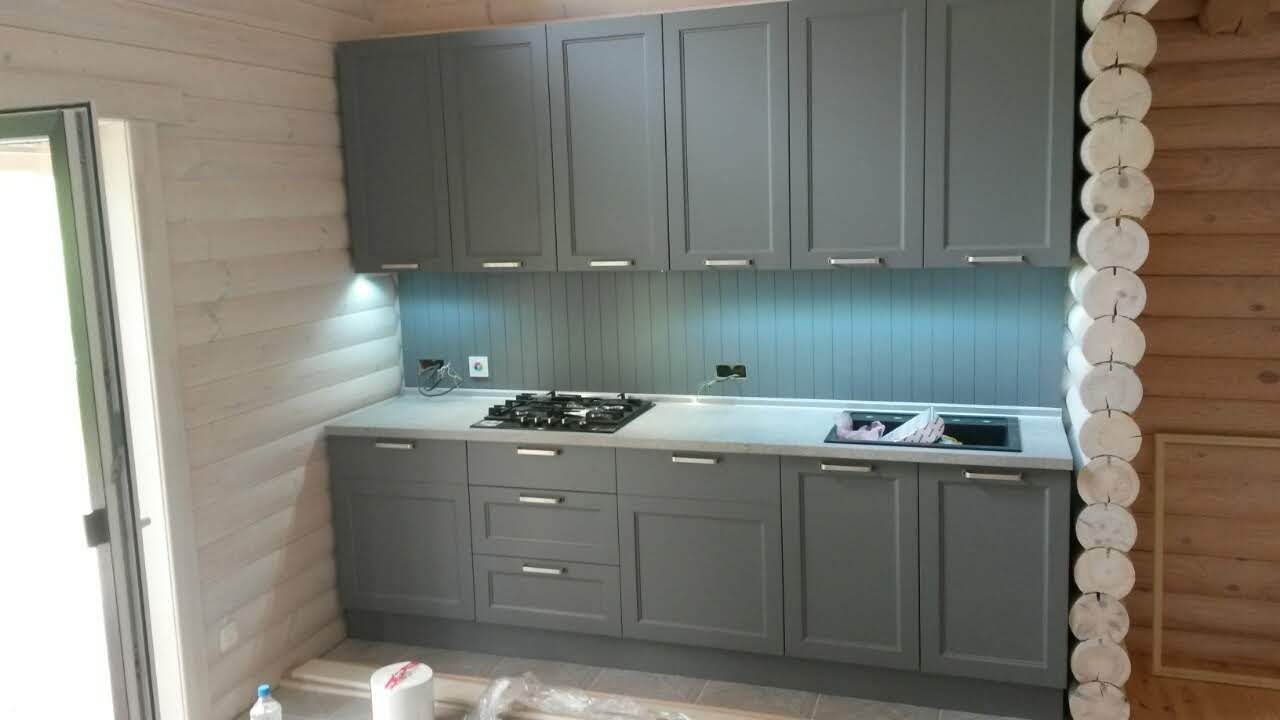 Szara zabudowa kuchenna w domu z bali, widoczne górne i dolne szafki z metalowymi uchwytami, płyta grzewcza i zlewozmywak, ściany wykończone drewnem.