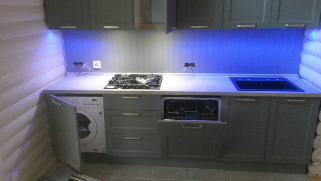 Nowoczesna szara zabudowa kuchenna w drewnianym domu z niebieskim oświetleniem LED podszafkowym; pralka i zmywarka ukryte za frontami.