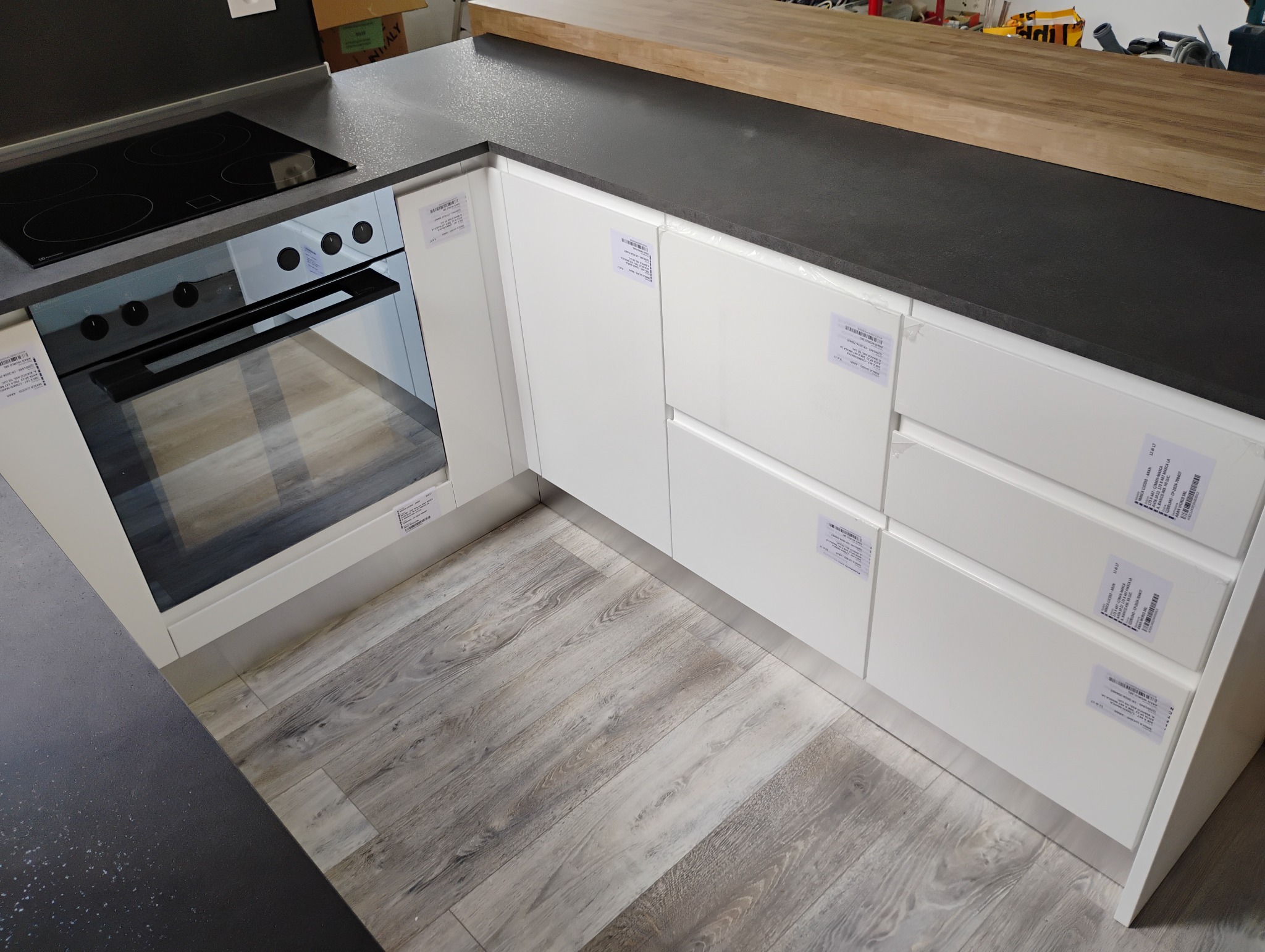 Fragment zabudowy kuchennej z białymi frontami szafek i szuflad oraz czarnym blatem, widoczny piekarnik elektryczny i płyta grzewcza, na podłodze panele imitujące drewno, na frontach szafek...