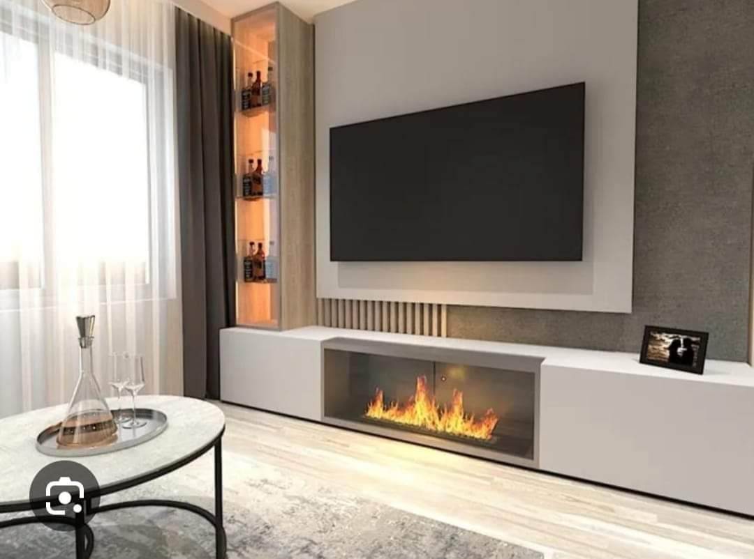 Nowoczesny salon z zabudową TV z biokominkiem, podświetlaną witryną na alkohol i okrągłym stolikiem kawowym z dekoracyjnym dzbankiem.