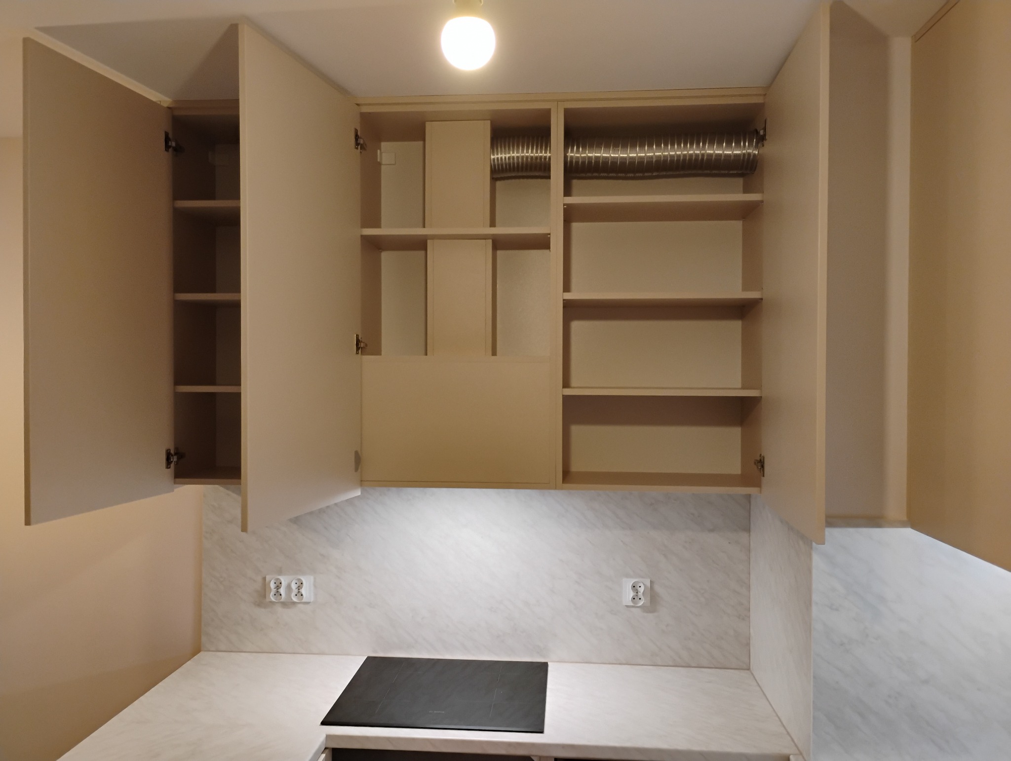 Beżowe szafki kuchenne wiszące, niektóre z otwartymi drzwiczkami, ukazujące puste półki i rurę wentylacyjną, nad blatem z płytą indukcyjną, oświetlone punktowym światłem.