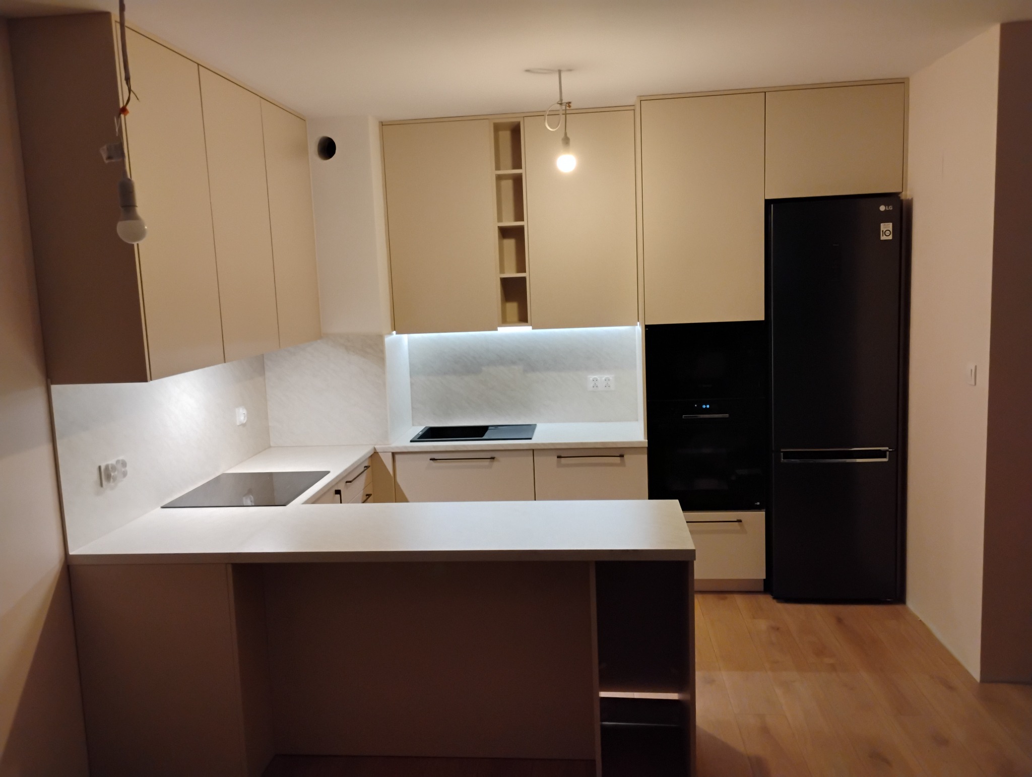 Nowoczesna kuchnia z beżowymi frontami, czarnym sprzętem AGD i wyspą kuchenną, oświetlenie LED pod szafkami, widoczna żarówka na kablu, drewniana podłoga.