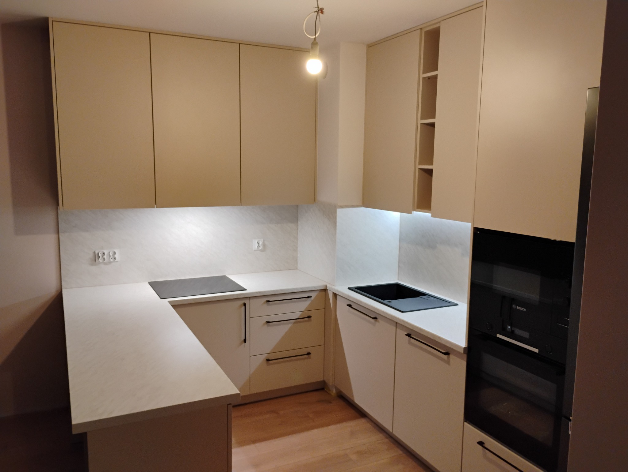 Jasna kuchnia z beżowymi szafkami, czarnym zlewem i płytą indukcyjną, oświetlona lampą wiszącą i światłem LED pod szafkami.