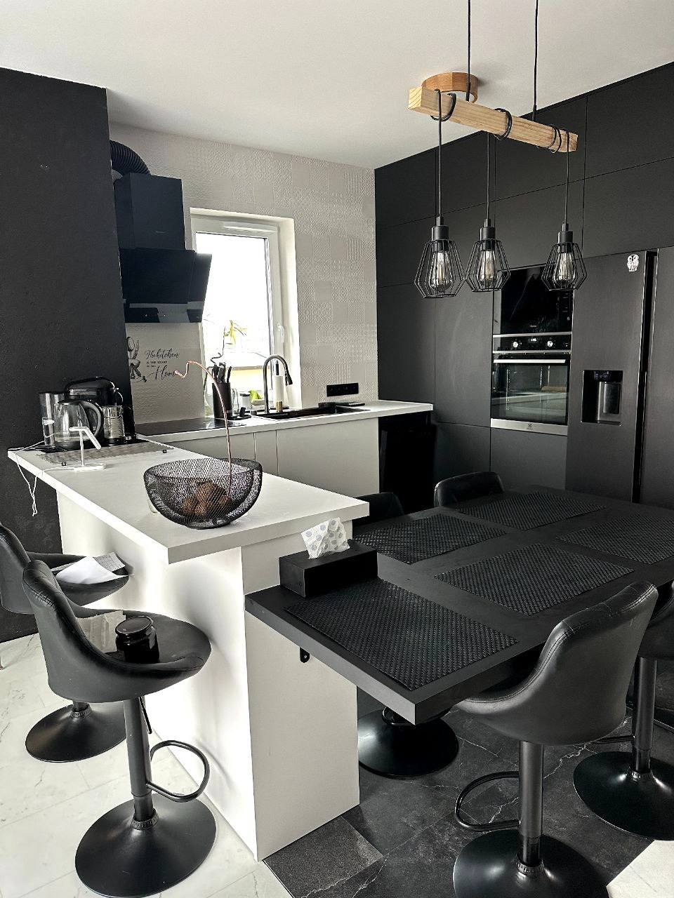 Nowoczesna kuchnia w czerni i bieli z wyspą i stołem barowym, oświetlona wiszącymi lampami z drewnianym elementem, z widocznym sprzętem AGD i dekoracyjnymi detalami.